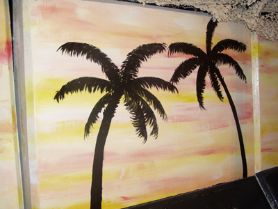 A pained mural on a concrete wall depicts palm trees against a yellow and pink sky.