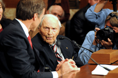 An elderly man and a younger man, both in suits, sit at a table behind microphones.  In the background are other people who are seated, and one man with a camera.
