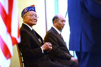 Two men in suits, one with a hat that indicates veteran status, are seated near an American flag.
