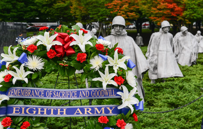 A wreath has ribbons with the words "Remembrance of Sacrifice and Goodwill" and "Eighth Army."  In the background are multiple statues of Soldiers who wear helmets and ponchos.