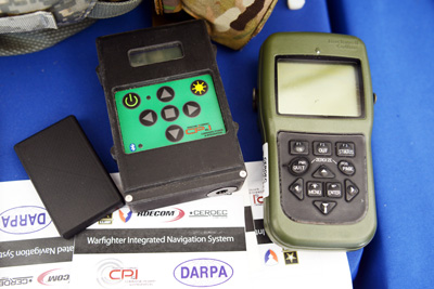 Two electronic devices sit on a table.  Paperwork says "DARPA" and "Warfighter Integrated Navigation System."